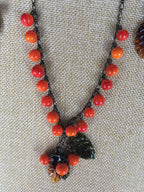 A glass beaded necklace with orange and red hues, featuring a unique pendant design. The necklace is displayed on a textured background.