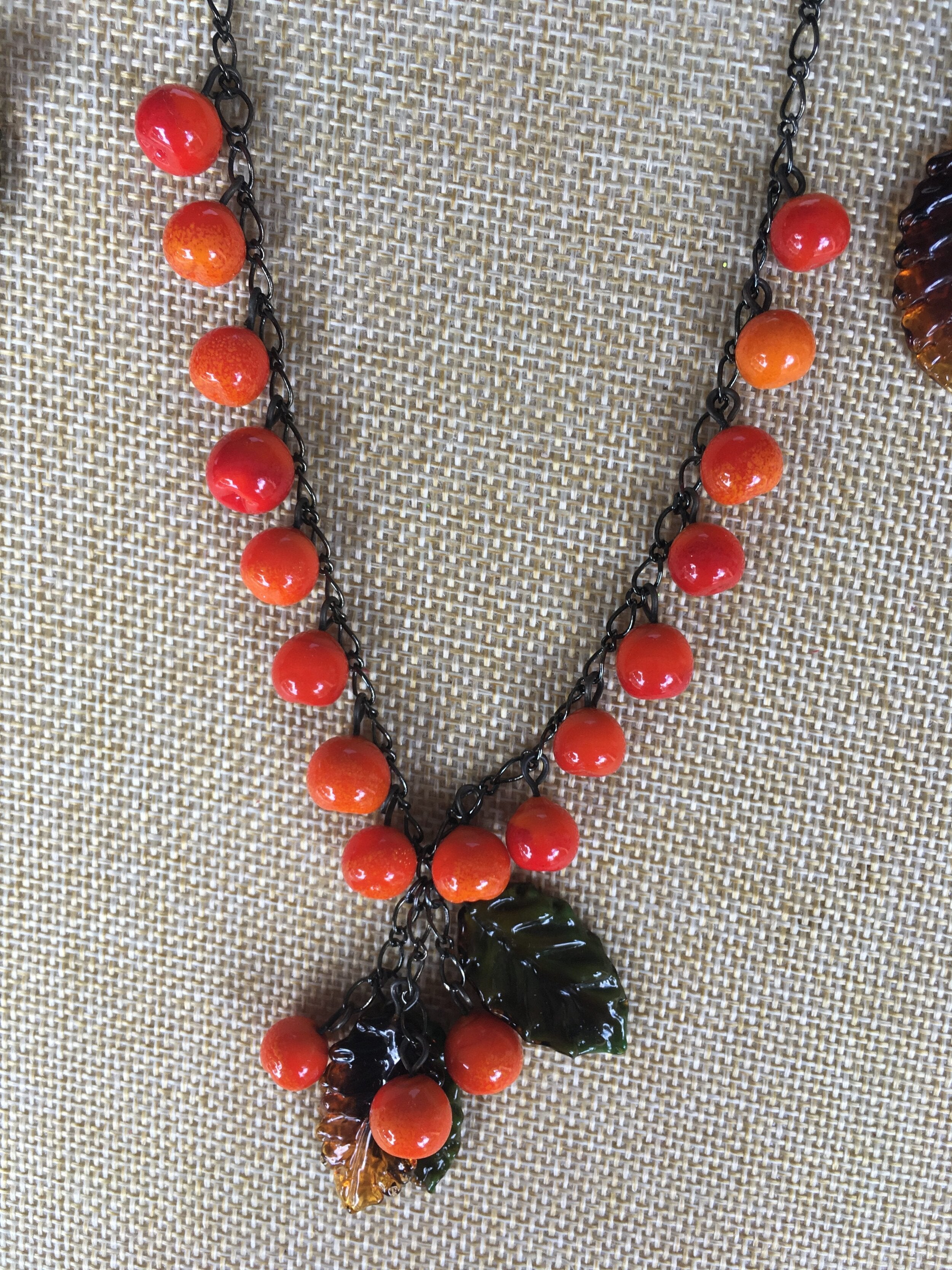 A glass beaded necklace with orange and red hues, featuring a unique pendant design. The necklace is displayed on a textured background.
