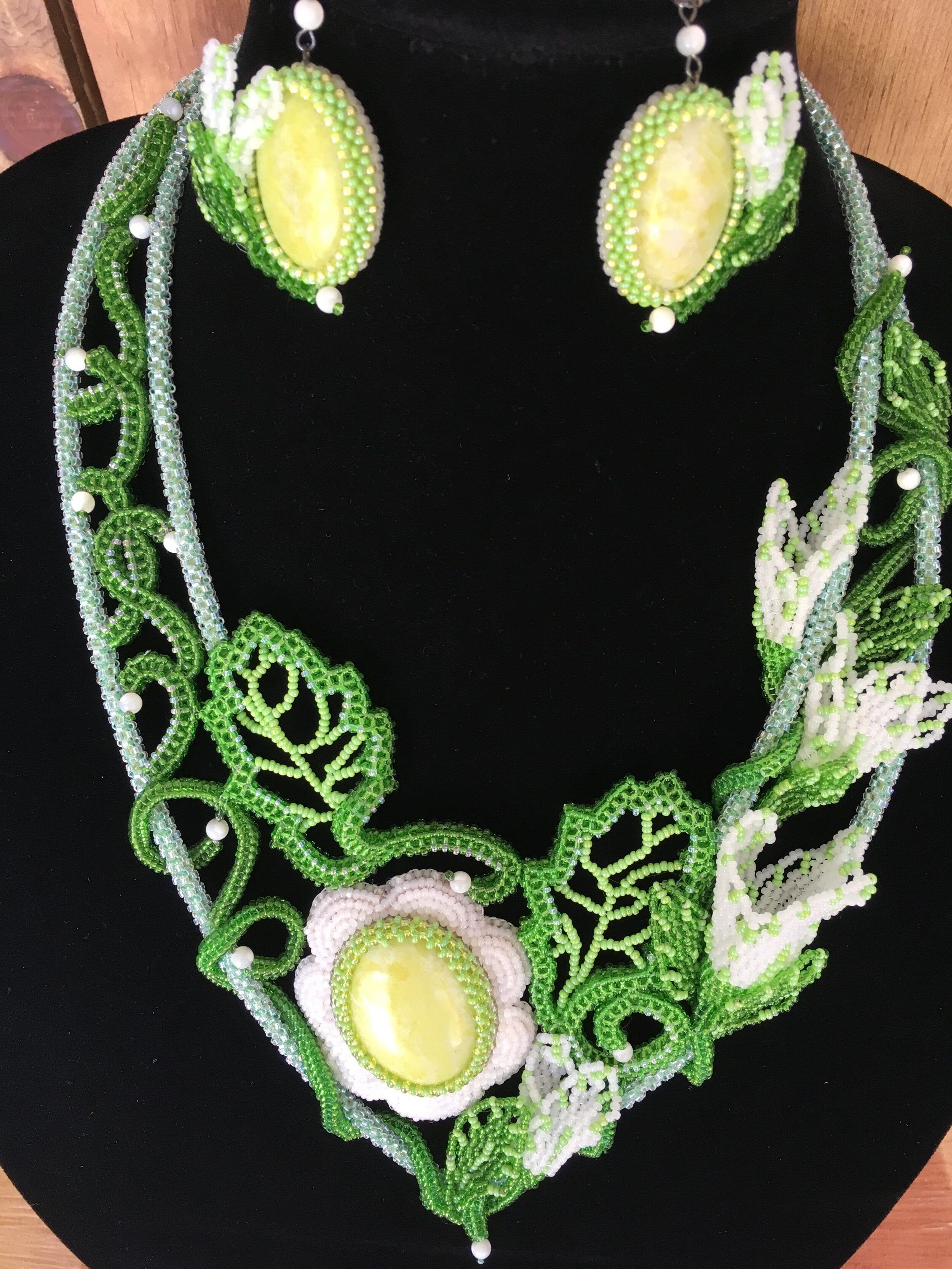 A necklace with a floral pattern, featuring green and white beads and accented with yellow jasper stones, displayed on a black stand.